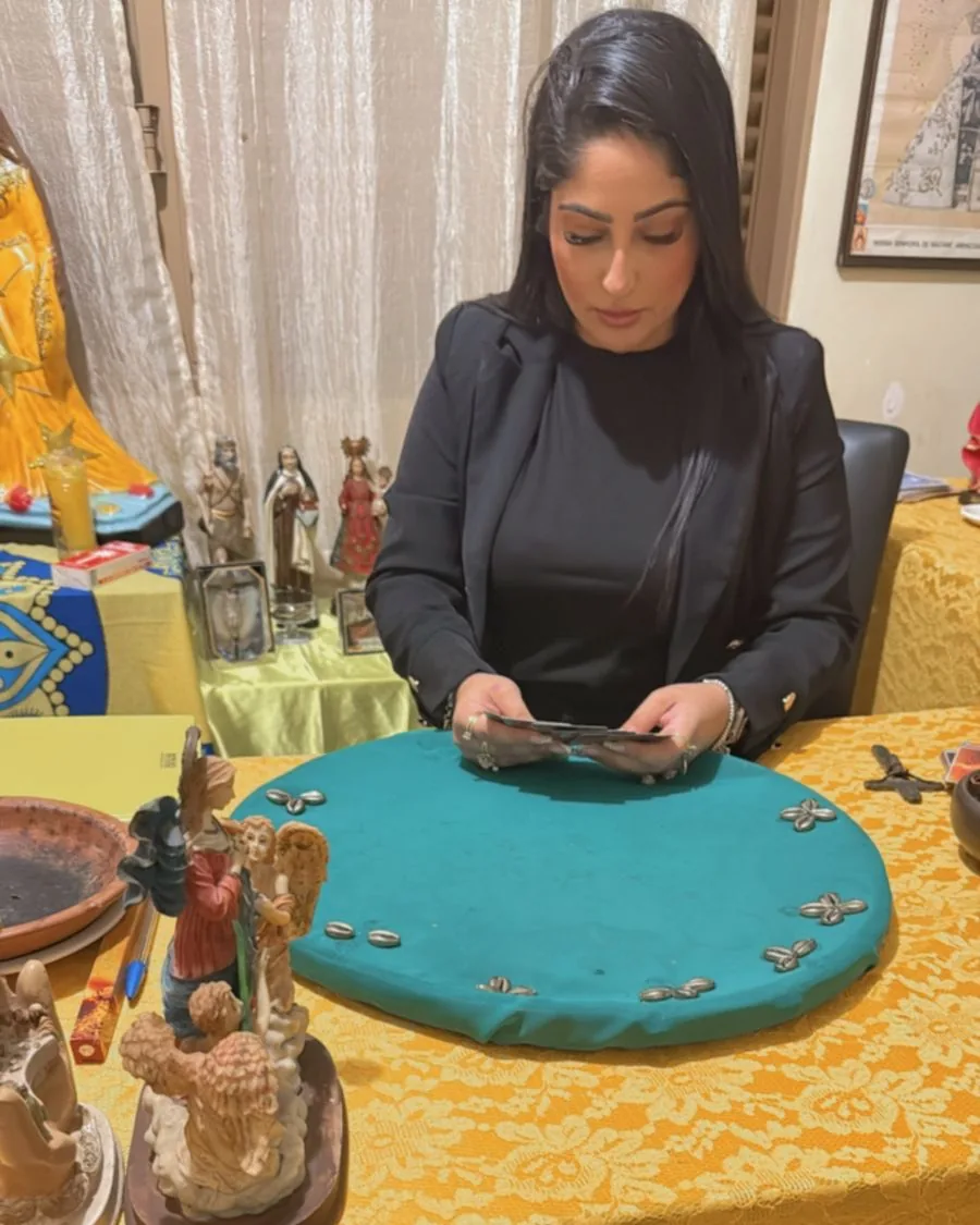Tarologa Day de blazer preto, sentada à mesa de atendimento com toalha de renda dourada, olhando as cartas que segura, com imagens de santos ao fundo